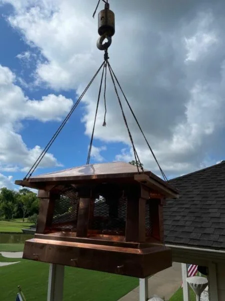 Copper chimney cap with mesh vents being hoisted for roof installation by Texas Chimney LLC