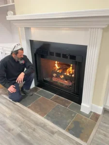 Man kneeling in front of white fireplace with burning logs, inspecting hearth area in residential home.