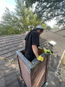 Technician in gray shirt repairing chimney on tiled roof with drill surrounded by trees