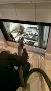 A technician using a long vacuum nozzle to clean the corners and floor of an empty fireplace firebox lined with light-colored brick panels.