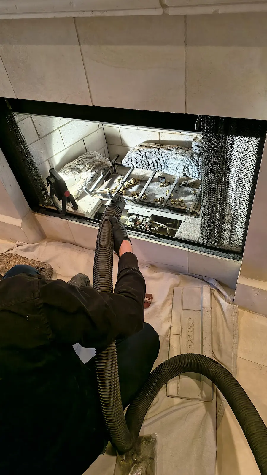 A technician using a long vacuum nozzle to clean the corners and floor of an empty fireplace firebox lined with light-colored brick panels.