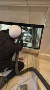 A side view of a technician kneeling on a drop cloth while using a heavy-duty vacuum hose to clean out the bottom of a built-in fireplace.