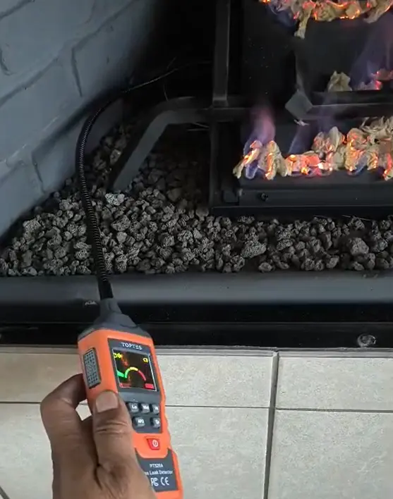 the individual is using a combustible gas leak detector (specifically a TopTES PT520A) to check for gas leaks around a lit gas fireplace
