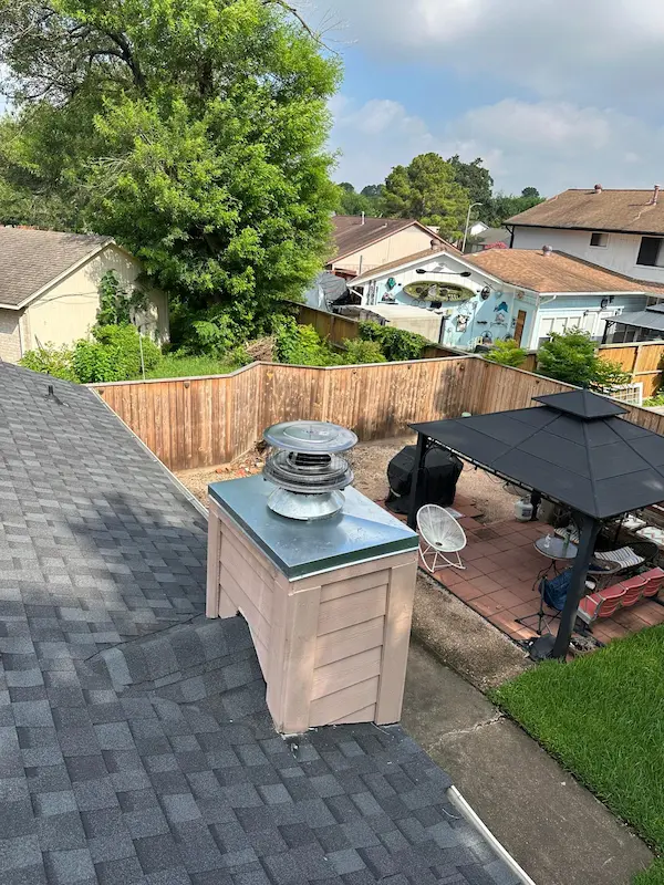 A wooden chimney chase with a newly installed metal chase pan and round rain cap on a dark shingled roof, overlooking a fenced residential backyard patio.